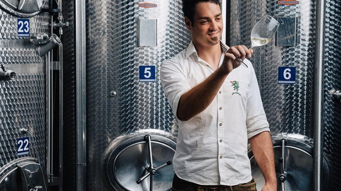 Ein junger Mann hält ein Weinglas und betrachtet den Inhalt. Im Hintergrund sind silberne Weinfässer zu sehen. | © Garber Patrick Weine