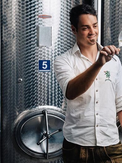 Ein junger Mann hält ein Weinglas und betrachtet den Inhalt. Im Hintergrund sind silberne Weinfässer zu sehen. | © Garber Patrick Weine