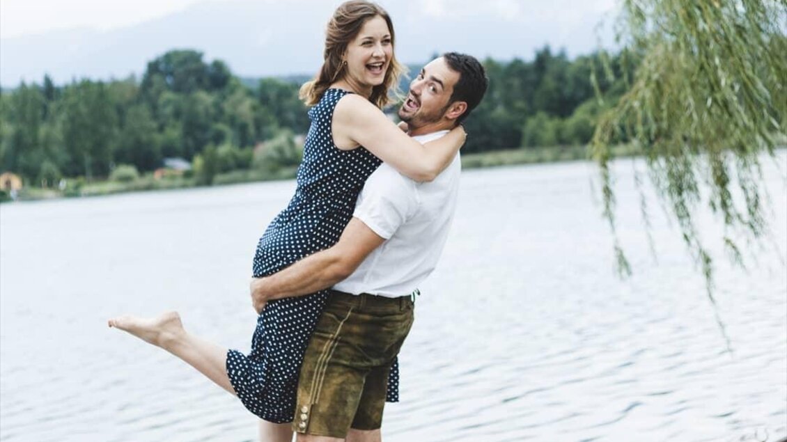 Ein glückliches Paar steht am Ufer eines Sees. Die Frau ist in einem blauen Kleid, der Mann trägt eine helle Bluse und schaut sie an. | © Gaber Patrick Weine