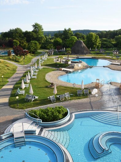 Ein schöner Aquapark mit mehreren Schwimmbecken und vielen Liegen. Umgeben von grünen Bäumen und einem klaren Himmel. | © Rogner Bad Blumau