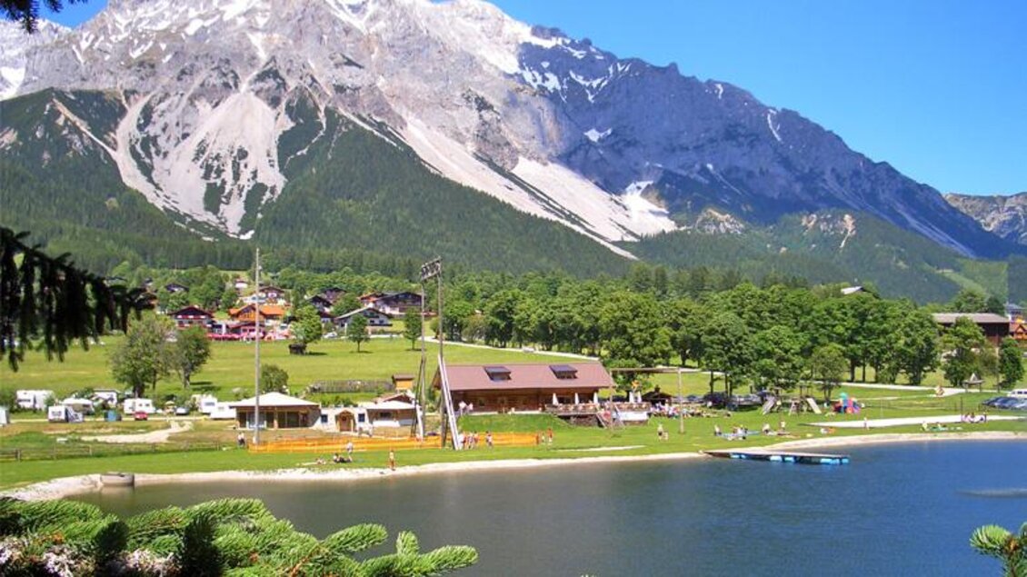Eine malerische Landschaft mit einem klaren See und grünen Wiesen. Im Hintergrund erhebt sich ein majestätischer Berg unter blauem Himmel. | © Photo Austria