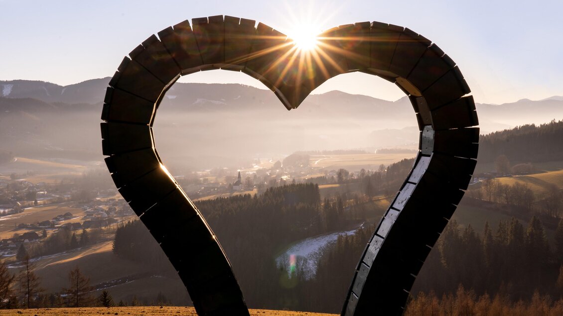 Ein Herz aus Holz, das in der Morgensonne strahlt. Im Hintergrund sind sanfte Hügel und eine malerische Landschaft zu sehen. | © Günther Breidler