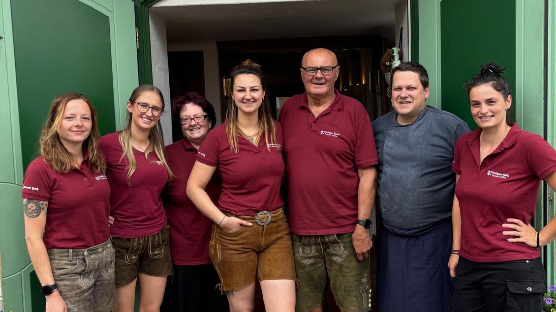 Eine Gruppe von sieben Personen steht vor einem Eingang mit offenen grünen Türen. Alle tragen einheitliche burgunderfarbene Shirts und lächeln in die Kamera. | © Gasthaus Ägydi