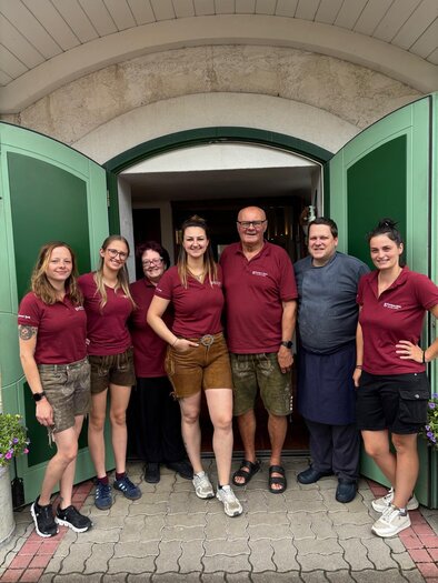 A group of seven people stands in front of an entrance with open green doors. They all wear matching burgundy shirts and smile at the camera. | © Gasthaus Ägydi