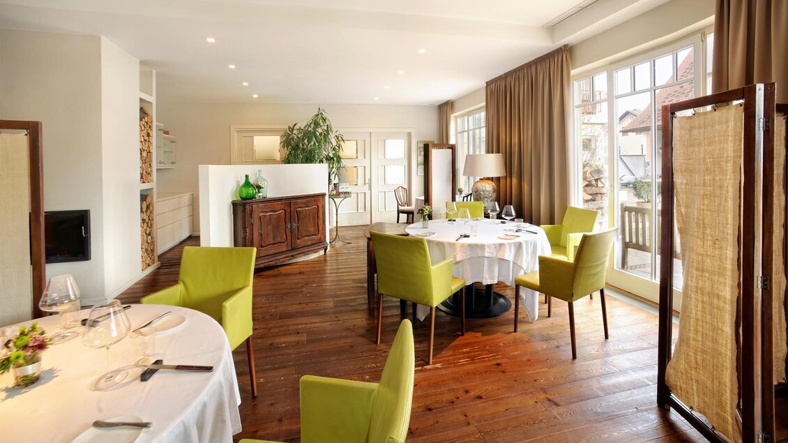 Ein elegantes Restaurant mit Holzböden und großen Fenstern. Die Tische sind mit weißen Tischdecken und modernen, grünen Stühlen gedeckt. | © Gasthaus Thaller
