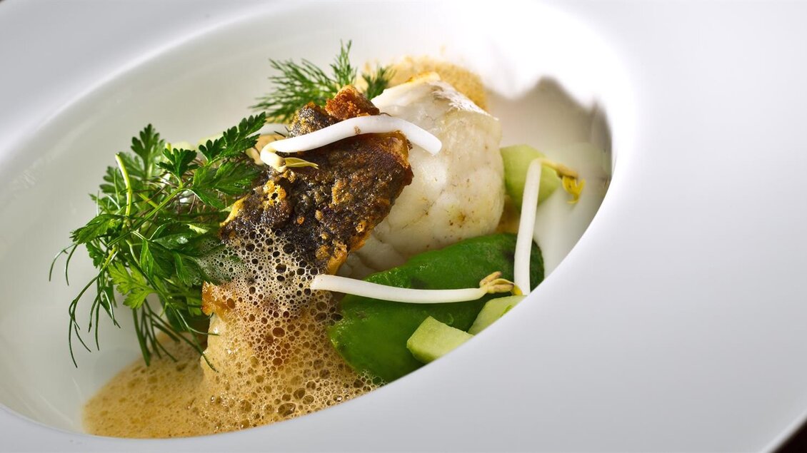 Ein kunstvoll angerichtetes Gericht mit Fisch, frischen Kräutern und einer aromatischen Brühe. Die Präsentation erfolgt in einer eleganten weißen Schale. | © Gasthaus Thaller