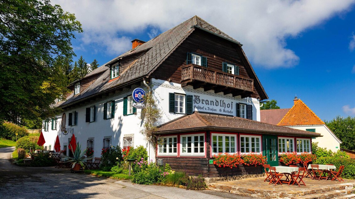 Ein charmantes Gasthaus mit einer traditionellen Holzfassade und einem blauen Himmel. Die Umgebung ist grün und einladend, mit Blumen und Sitzmöglichkeiten im Freien. | © TV Region Graz - Harry Schiffer