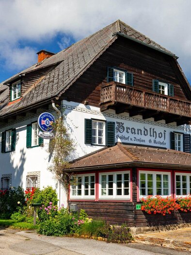 Ein charmantes Gasthaus mit einer traditionellen Holzfassade und einem blauen Himmel. Die Umgebung ist grün und einladend, mit Blumen und Sitzmöglichkeiten im Freien. | © TV Region Graz - Harry Schiffer