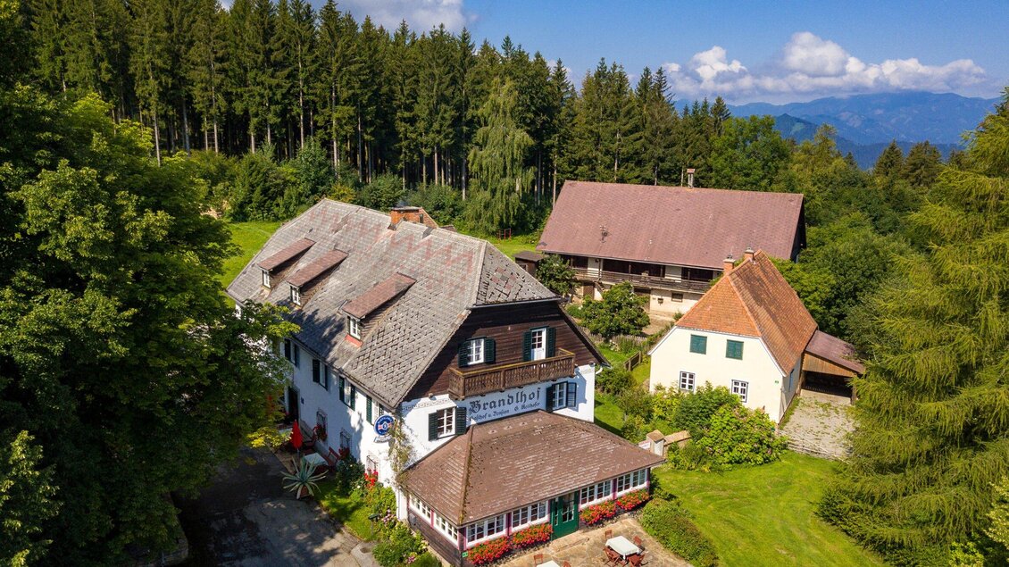Ein malerisches Landhaus mit verschiedenen Nebengebäuden, umgeben von grünen Bäumen und Bergen im Hintergrund. Die Szene strahlt eine ruhige und einladende Atmosphäre aus. | © TV Region Graz - Harry Schiffer