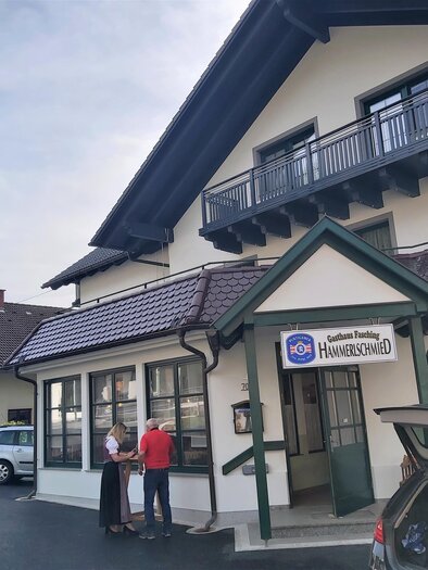 Ein charmantes Gasthaus mit einem einladenden Eingangsbereich. Zwei Personen stehen vor dem Gebäude und unterhalten sich.