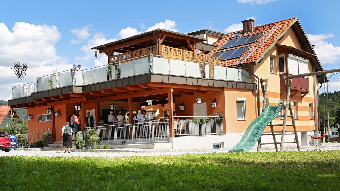 Ein modernes Haus mit einem großen Balkon und einer grünen Rutsche im Garten. Es sind mehrere Personen im Freien versammelt. | © TV Region Graz - Lunghammer