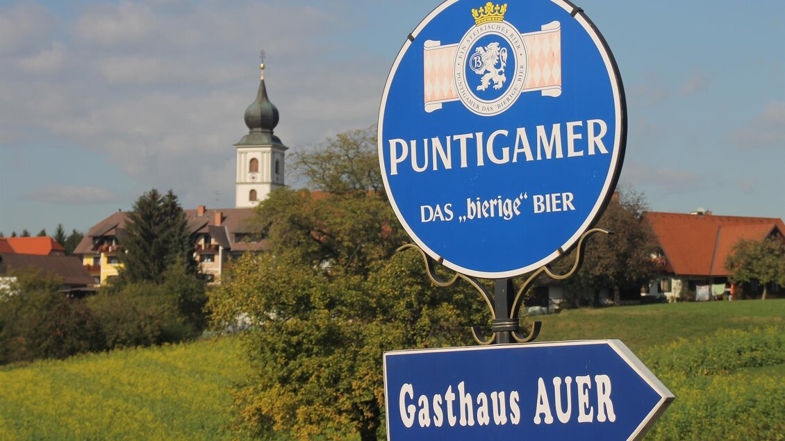 Ein Schild für Puntigamer Bier mit dem Hinweis auf das Gasthaus Auer. Im Hintergrund ist eine Kirche und eine grüne Landschaft zu sehen. | © Gasthaus Auer