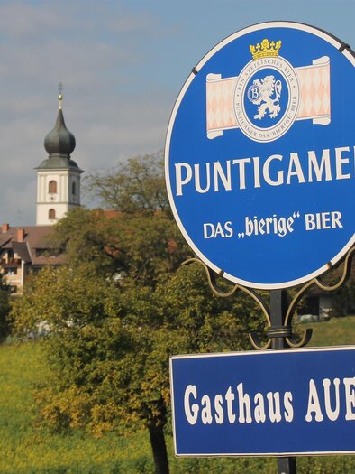 A sign for Puntigamer beer with a reference to the Auer inn. In the background, a church and a green landscape can be seen. | © Gasthaus Auer