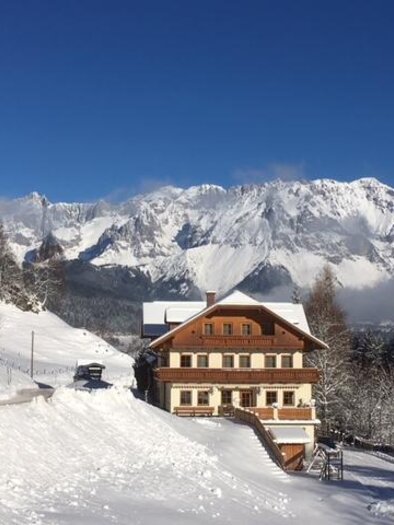 Eine malerische Winterlandschaft mit schneebedeckten Bergen. Im Vordergrund steht ein traditionsreiches Holzhaus. | © Bankwirt