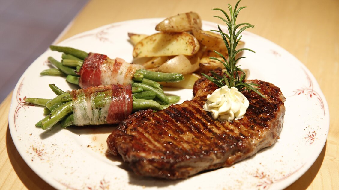 Ein saftiges Steak mit gegrillten Bohnen im Speckmantel und knusprigen Kartoffeln. Das Gericht ist dekoriert mit einem Rosmarinzweig und einem Klecks Butter. | © Herbert Raffalt