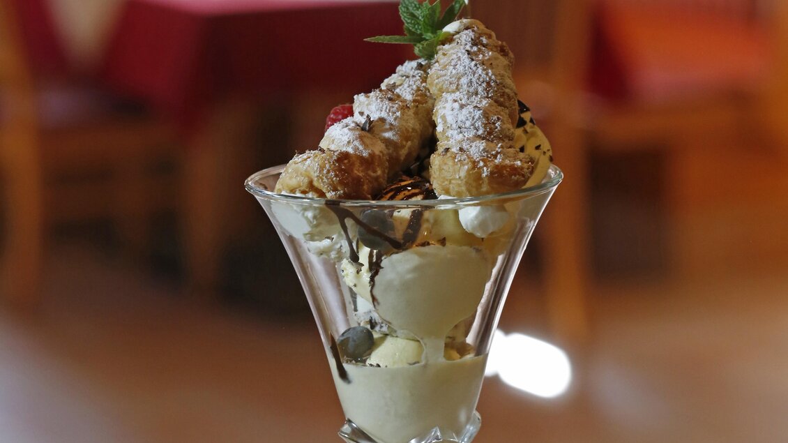 Ein köstliches Eisbecher mit mehreren Sorten Eis, Sahne und einem dekorativen Topping. Im Hintergrund sind verschwommene Tische und Stühle in einem Restaurant zu sehen. | © Herbert Raffalt