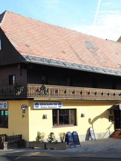 Ein charmantes, zweigeschossiges Gebäude mit einem rot gedeckten Dach. Die Fassade ist gelb, und es gibt einen Balkon sowie ein Restaurantzeichen. | © Steirische Rucksackdörfer
