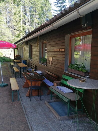 Ein gemütliches Holzhaus mit Tischen und Stühlen im Freien. Neben dem Gebäude steht ein Sonnenschirm und umgeben ist es von Bäumen. | © Gasthaus Fuchsbau