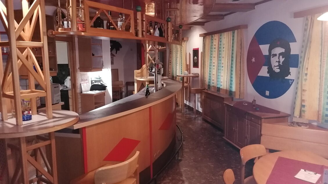 Ein gemütliches Restaurant mit einer großen Bar und Holzdekor. An der Wand ist ein großes Wandbild eines Mannes und die Fenster sind mit bunten Vorhängen geschmückt. | © Gasthaus Fuchsbau