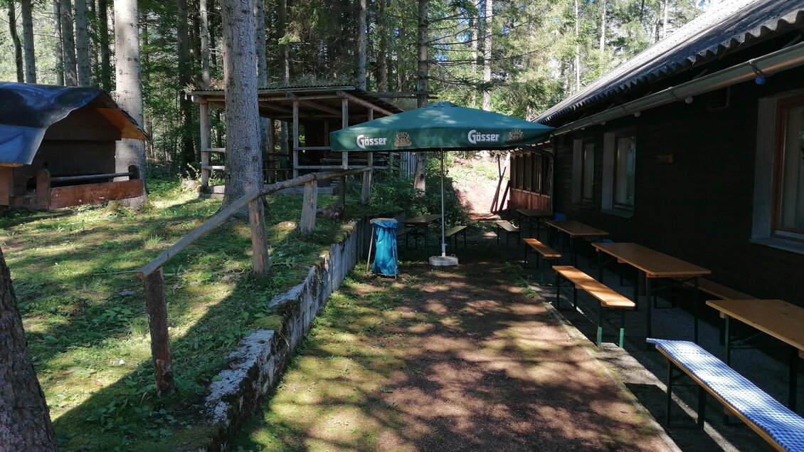 Ein gemütlicher Außenbereich mit Tischen und Bänken im Wald. Über einem der Tische steht ein Sonnenschirm. | © Gasthaus Fuchsbau