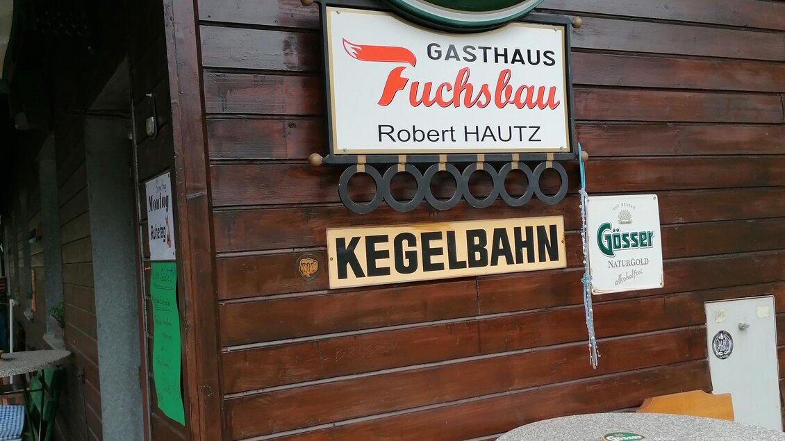 Eine Katze sitzt auf einem Stuhl vor einem Holzgebäude mit verschiedenen Schildern. Das Schild zeigt das Logo von Gösser und Namen des Gasthauses. | © Gasthaus Fuchsbau