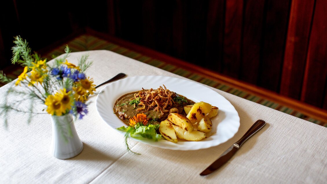 Ein schön angerichteter Teller mit Fleisch, knusprigen Zwiebeln und Bratkartoffeln. Daneben steht eine kleine Vase mit bunten Blumen auf dem Tisch. | © Tourismusverband Murau