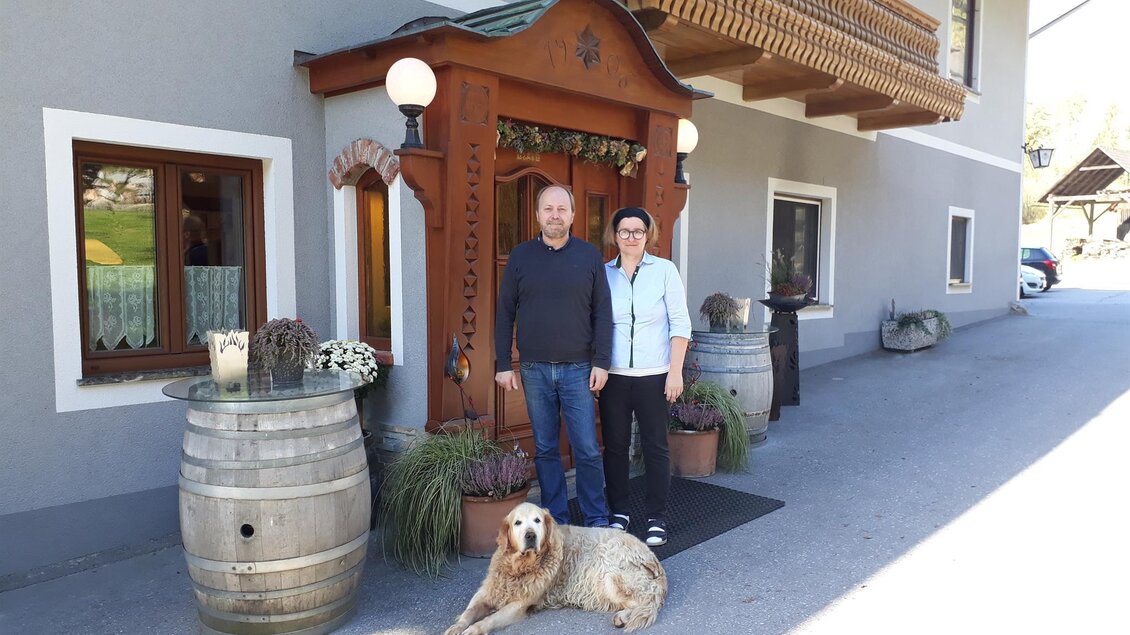 Ein freundliches Pärchen steht vor einem traditionellen Gebäude. Ein Hund liegt entspannt neben ihnen. | © TV Lipizzanerheimat