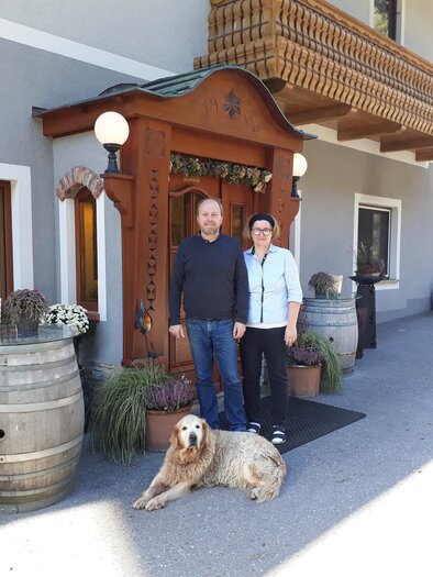 Ein freundliches Pärchen steht vor einem traditionellen Gebäude. Ein Hund liegt entspannt neben ihnen. | © TV Lipizzanerheimat