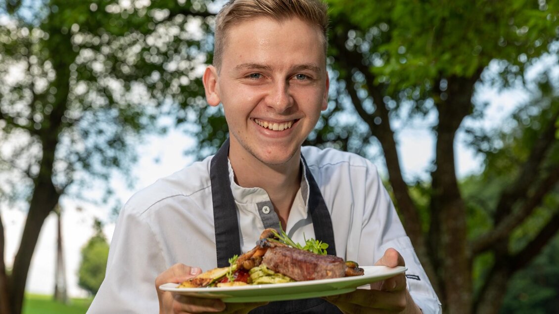 Ein junger Mann lächelt, während er einen Teller mit gegrilltem Fleisch und frischem Gemüse hält. Im Hintergrund sind grüne Bäume und ein klarer Himmel zu sehen. | © TV Region Graz - Harry Schiffer