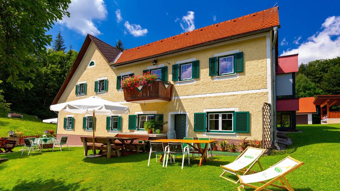 Ein charmantes Haus mit grünen Fensterläden in einer sonnigen Umgebung. Im Vordergrund befinden sich Tische, Stühle und Liegen auf einer grünen Wiese. | © TV Region Graz - Harry Schiffer