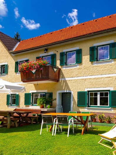 Ein charmantes Haus mit grünen Fensterläden in einer sonnigen Umgebung. Im Vordergrund befinden sich Tische, Stühle und Liegen auf einer grünen Wiese. | © TV Region Graz - Harry Schiffer