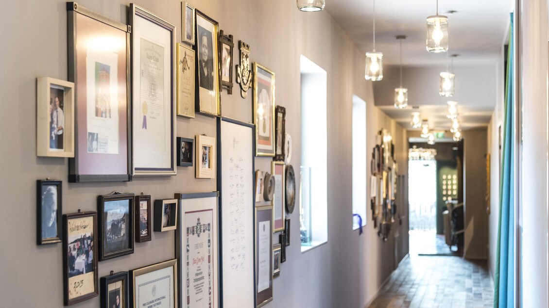 Ein heller Flur mit zahlreichen gerahmten Bildern und Auszeichnungen an der Wand. Die Decke ist mit modernen Lampen beleuchtet. | © Werner Krug