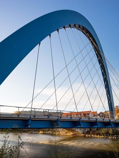 A modern blue bridge with an elegant arch structure over a river. In the background, buildings and trees can be seen. | © M. Meieregger