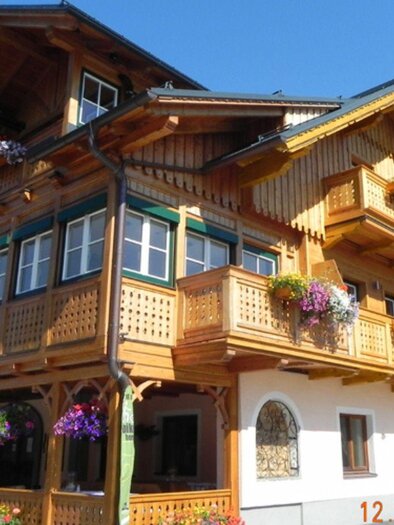 A traditional alpine wooden house with many flower balconies. The facade is bright and welcoming, surrounded by clear skies. | © Landhotel-Restaurant Kanzler