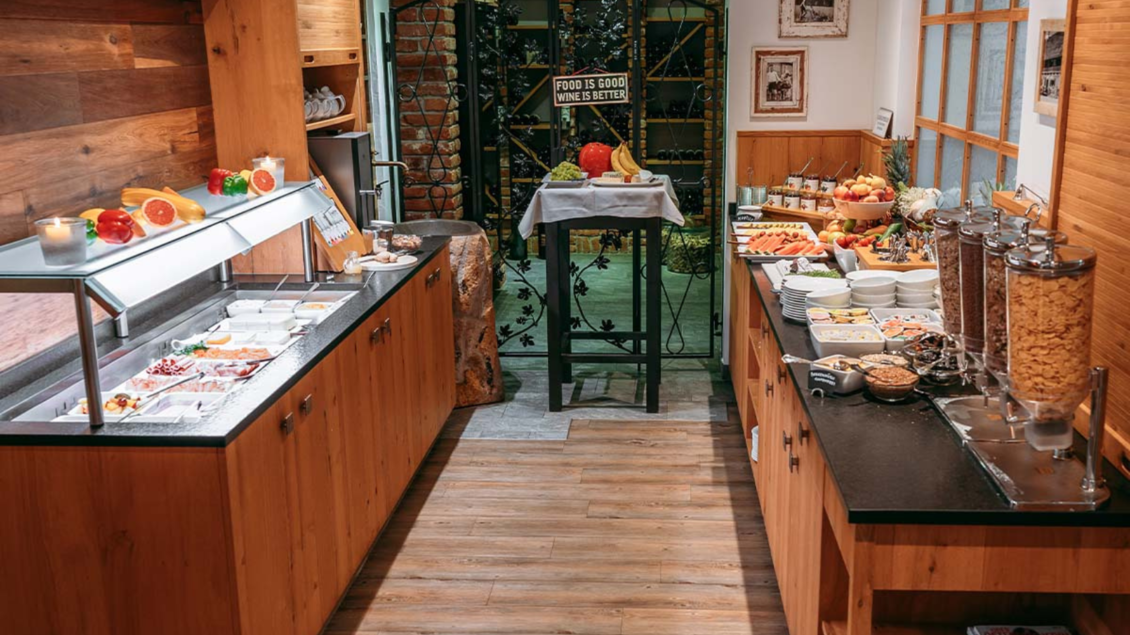 Ein einladender Frühstücksraum mit Holzregalen und einer Auswahl an frischem Obst, Müsli und Aufschnitt. Auf einem Tisch stehen weitere Speisen bereit, um den Morgen zu genießen. | © Gasthaus Kanzler