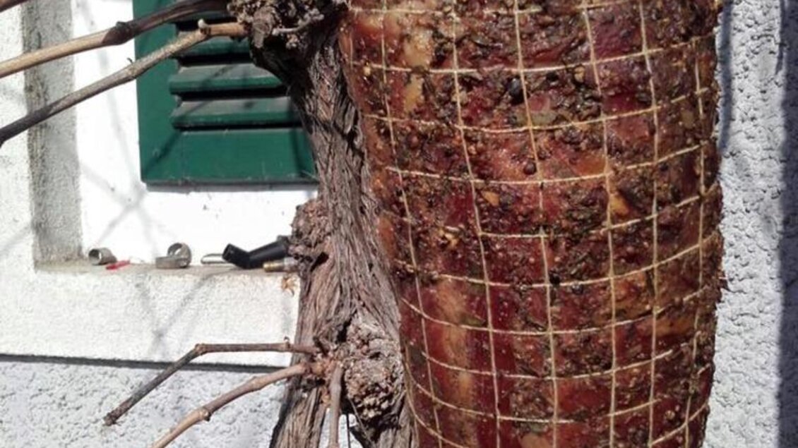 Ein Stück Schinken ist an einer Wand aufgehängt und mit einem Netz umwickelt. Im Hintergrund sind einige Weinstöcke zu sehen. | © Familie Koschak