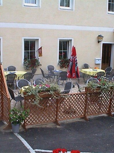 An inviting terrace with yellow tables and chairs, surrounded by flowers. In the background, there is a bright building with windows. | © Gasthaus Krapinger