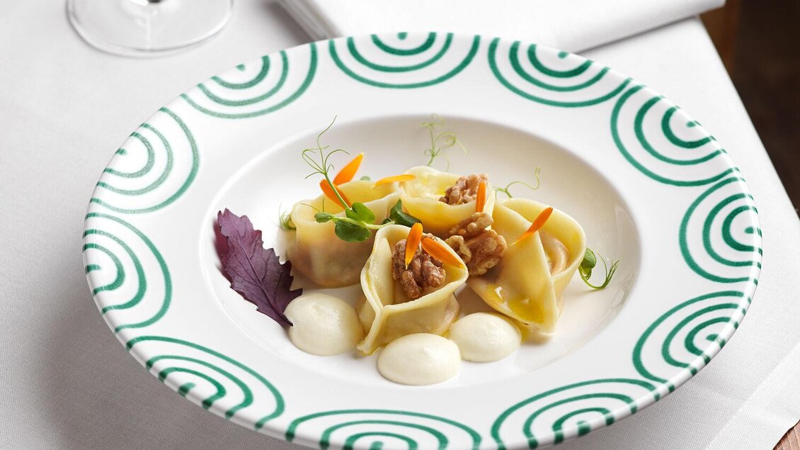 Ein kunstvoll angerichteter Teller mit gefüllten Teigtaschen, garniert mit buntem Gemüse. Daneben steht ein Glas Weißwein auf einem eleganten Tisch. | © TAUROA GmbH