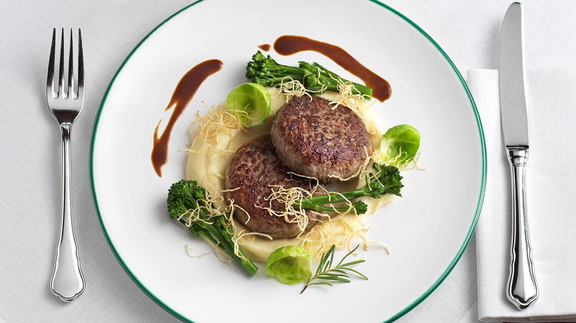 Ein elegantes Gericht mit zwei saftigen Rindersteaks auf einer Bett aus Lauch und frischem Gemüse. Dazu ein Glas roten Wein auf einem weißen Tisch. | © TAUROA GmbH