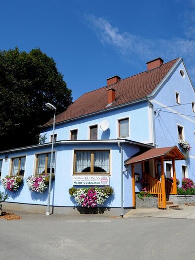 Ein blaues Gebäude mit einem roten Dach und bunten Blumen an den Fenstern. Darüber ist ein klarer blauer Himmel mit einigen Wolken. | © A. Steininger