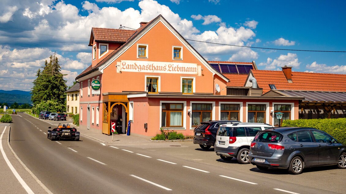 Ein gemütliches Gasthaus mit einer orangefarbenen Fassade, umgeben von Autos und einem klaren Himmel. Die Straße führt direkt am Gebäude vorbei. | © Wolfgang Speckner