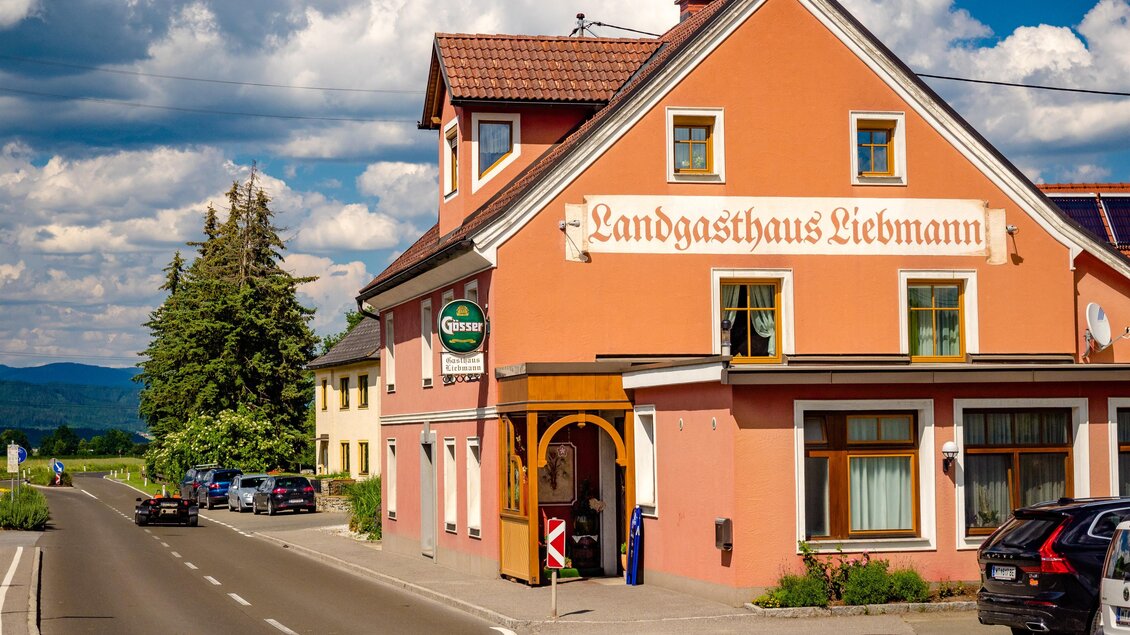 Ein charmantes Gasthaus mit einer orangefarbenen Fassade steht entlang einer Straße. Im Hintergrund sind grüne Bäume und der Himmel mit einigen Wolken zu sehen. | © Wolfgang Speckner