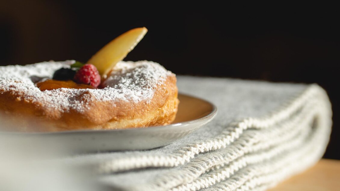 Ein köstlicher Donut mit Puderzucker und frischen Früchten auf einem Teller. Der Hintergrund ist weich und unscharf, was das Gebäck hervorhebt. | © © 2022 Alwin Strasser - strasserconcept