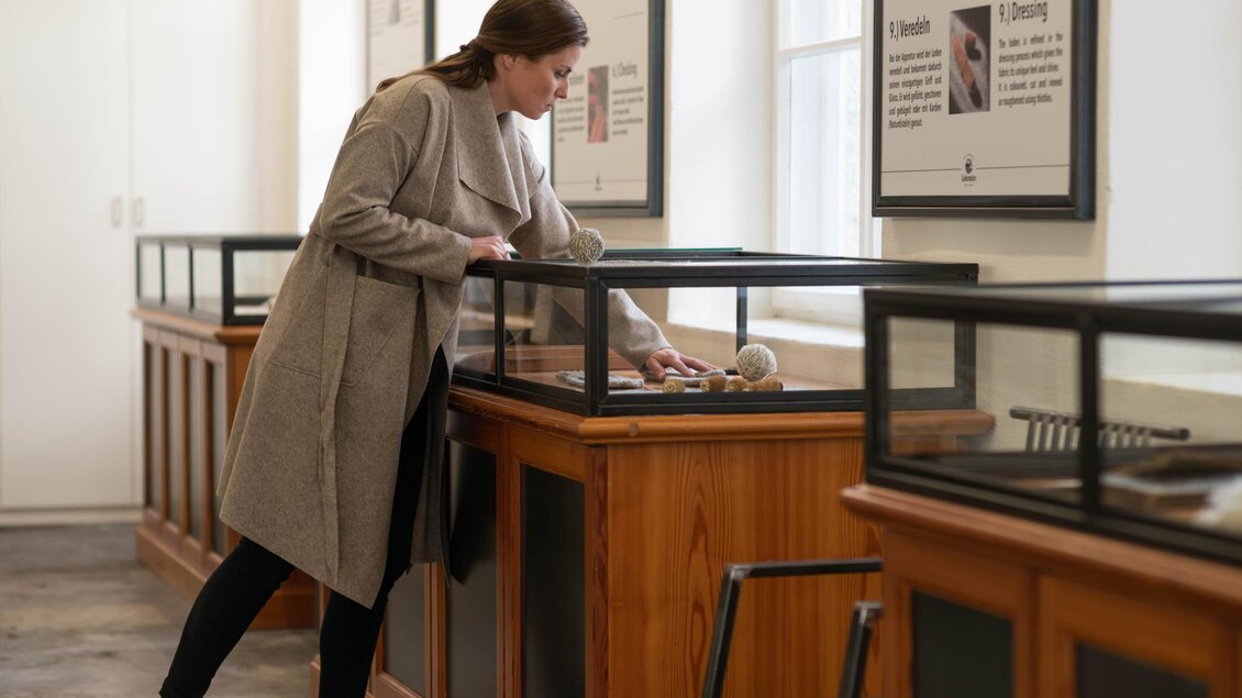 Eine Frau untersucht eine Ausstellung in einem Museum. Sie steht vor einem Vitrinenschrank und schaut sich die Objekte an. | © Lodenwalker Ramsau
