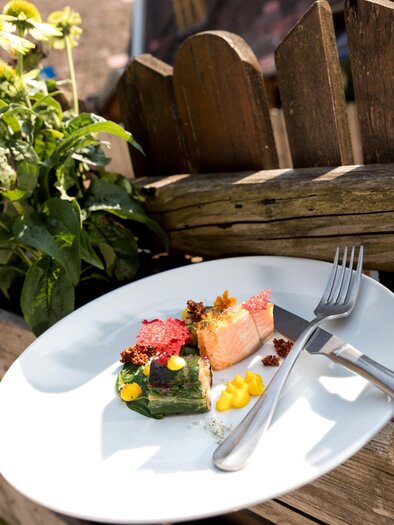 A plate with colorful food, consisting of various pieces of vegetables and fruits, is placed on an old wooden fence. In the background, green plants and flowers can be seen. | © Region Graz - Harry Schiffer