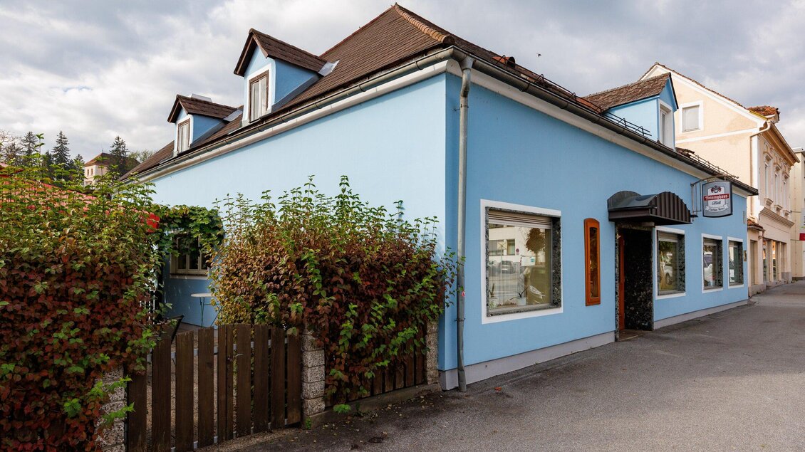 Ein blaues Haus an einer Straße mit kleinen Fenstern und einer umgebenden Hecke. Der Himmel ist bewölkt und es gibt eine ruhige Atmosphäre. | © TV Region Graz - Harry Schiffer