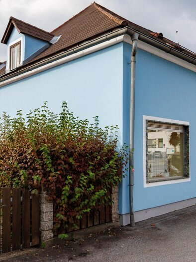 Ein blaues Haus an einer Straße mit kleinen Fenstern und einer umgebenden Hecke. Der Himmel ist bewölkt und es gibt eine ruhige Atmosphäre. | © TV Region Graz - Harry Schiffer