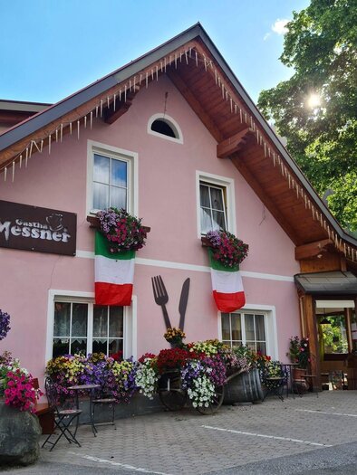 Ein charmantes Restaurant mit einer rosa Fassade und bunten Blumenkübeln. Flaggen und einladende Details sorgen für eine freundliche Atmosphäre. | © Gasthaus Messner