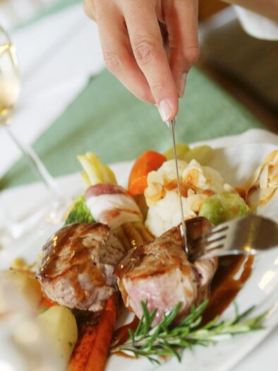 A plate with braised meat, vegetables, and a fine sauce. A hand is cutting the food while a glass of white wine stands in the background. | © TVB Thermen & Vulkanland