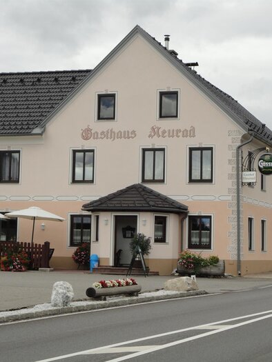 Ein gemütliches Gasthaus mit weißer Fassade und einem schwarzen Dach. Vor dem Gebäude stehen Tische mit Sonnenschirmen und Blumenarrangements. | © Gasthaus Neurad